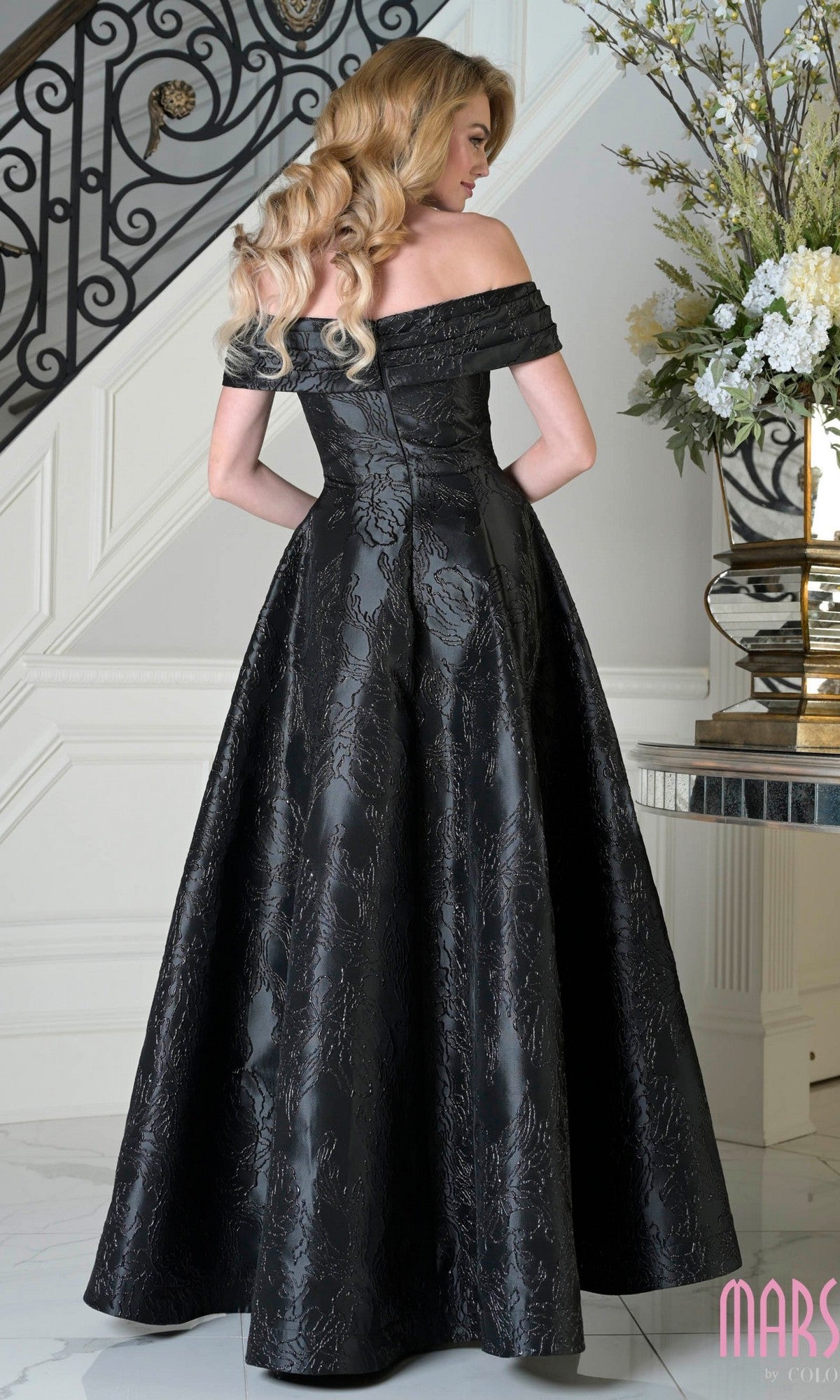 A woman in the Marsoni MV1395 off-the-shoulder jacquard long formal dress stands by a staircase and floral arrangement, facing away from the camera.